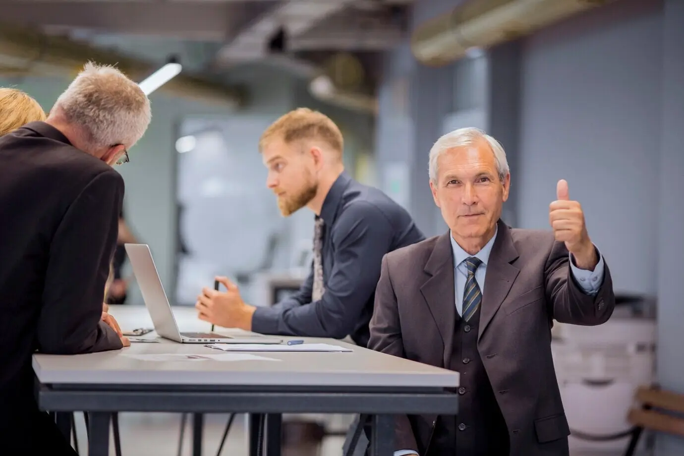 Ein leitender Geschäftsmann sitzt vor seinem im Büro diskutierenden Team und zeigt den Daumen hoch.